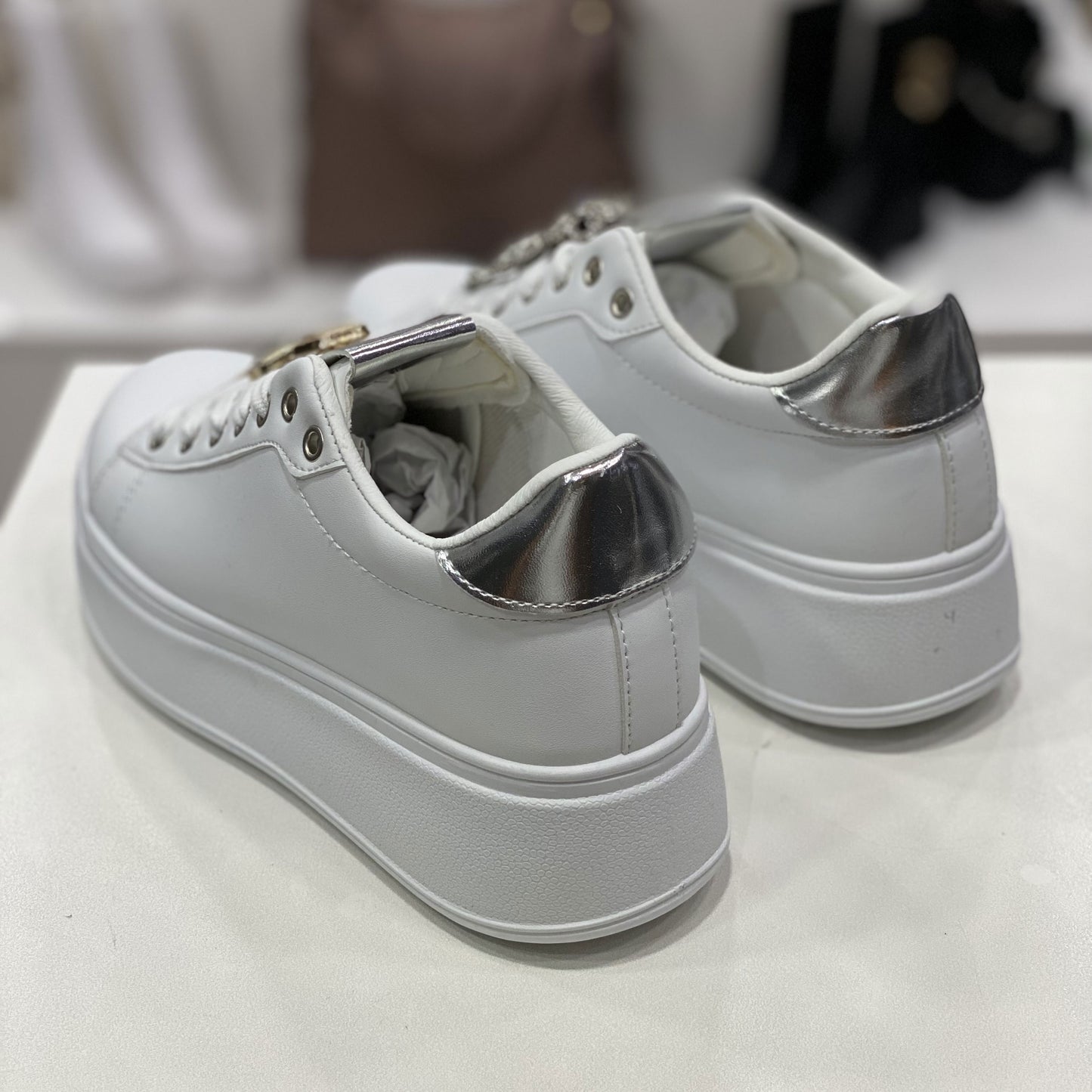 Un paio di scarpe da ginnastica bianche con dettagli metallici argentati sul retro. La suola alta e piatta conferisce un aspetto moderno e di tendenza, perfetto per un look casual ma ricercato.