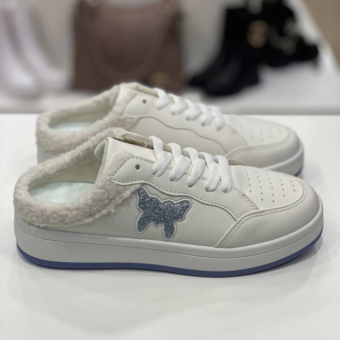 Sneakers a ciabatta bianche con decorazione a forma di farfalla glitterata e interno in peluche nero, vista laterale su sfondo neutro