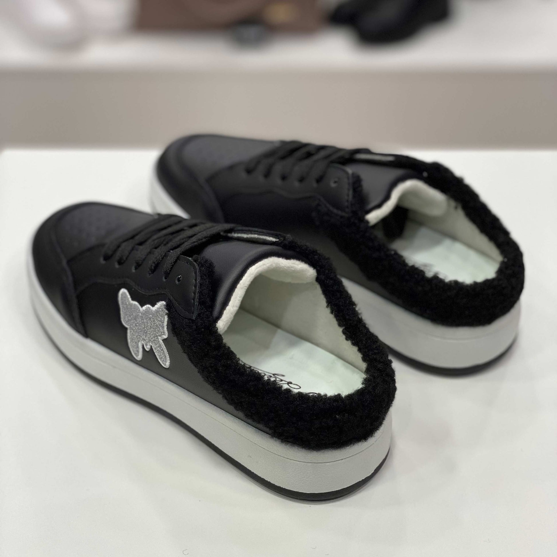 Parte posteriore di sneakers a ciabatta nere con interno peluche e suola bianca, vista del tallone su sfondo neutro