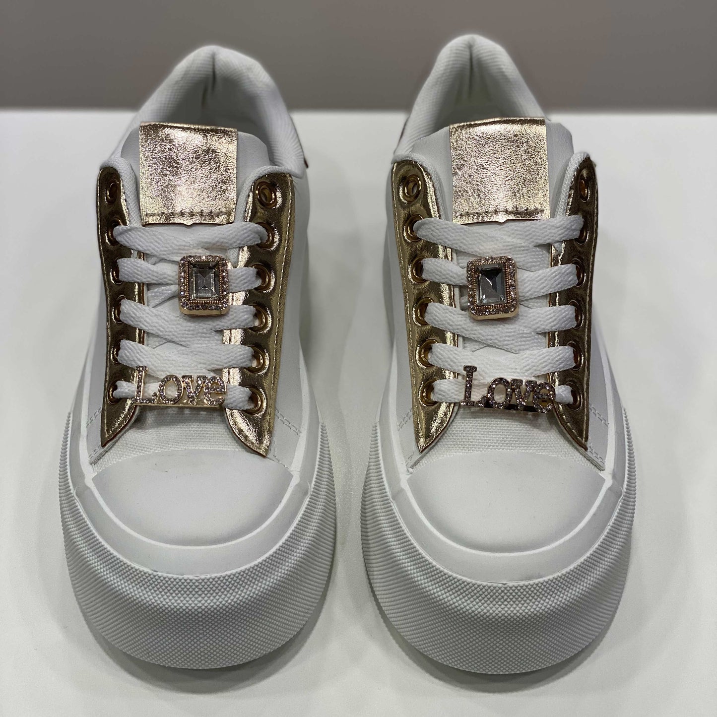 Sneakers bianca con dettagli dorati e decorazioni gioiello sui lacci, vista frontale su sfondo neutro