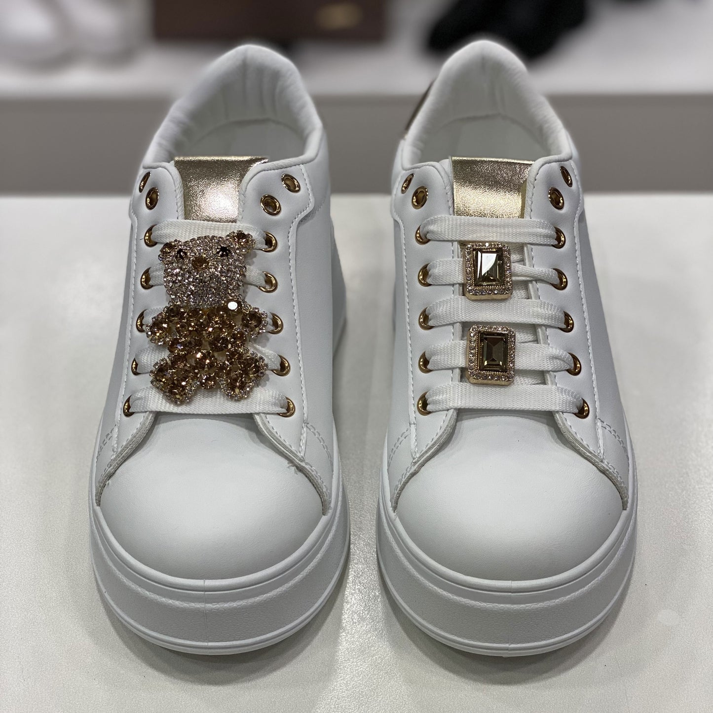 Scarpe sportive bianche con linguetta oro e con decorazioni uniche sui lacci: una scarpa ha un orsetto luccicante, mentre l'altra è adornata con due fibbie quadrate con pietre nere. Un tocco di personalità e stile glamour per chi cerca accessori distintivi.