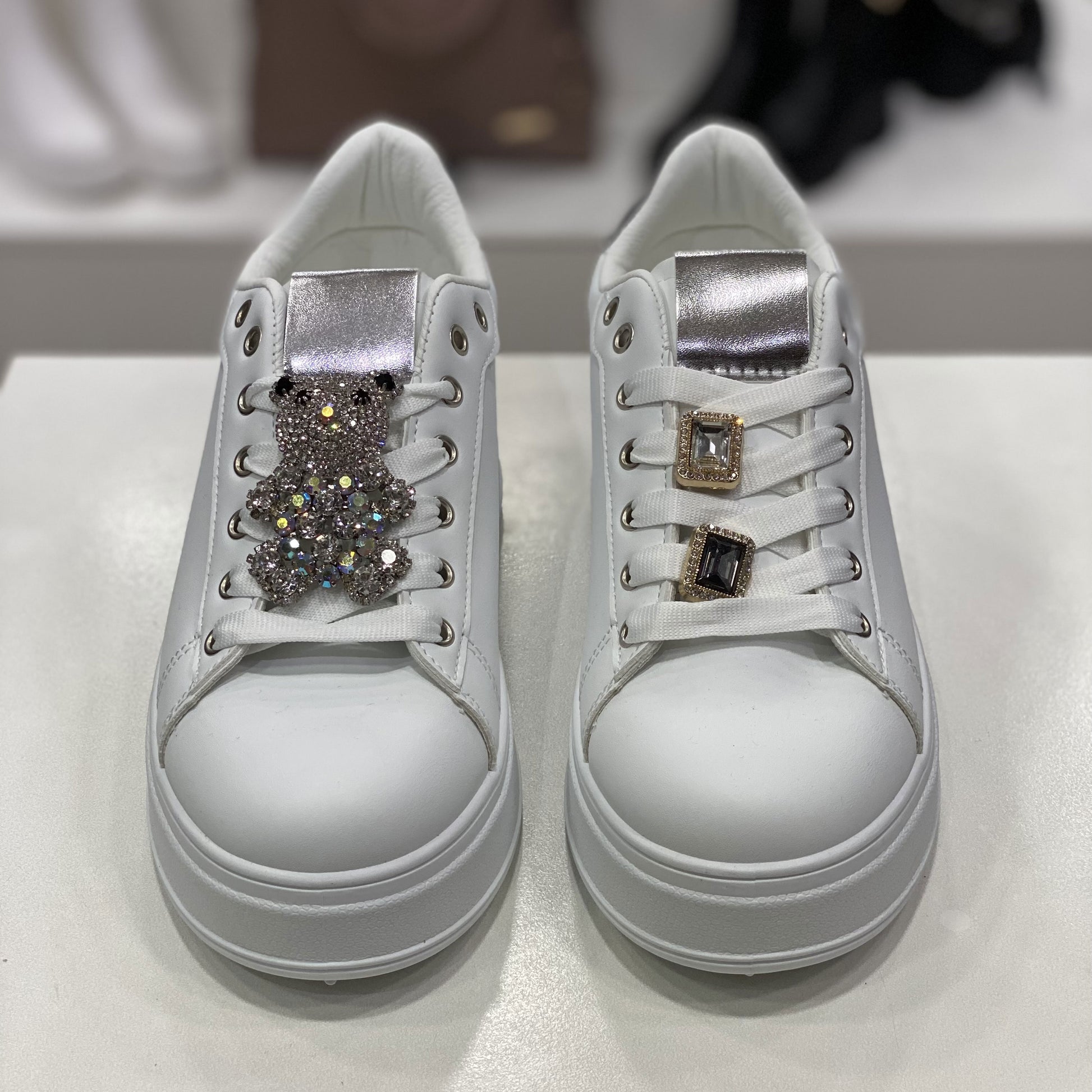 Scarpe sportive bianche con decorazioni uniche sui lacci: una scarpa ha un orsetto luccicante, mentre l'altra è adornata con due fibbie quadrate con pietre nere. Un tocco di personalità e stile glamour per chi cerca accessori distintivi.