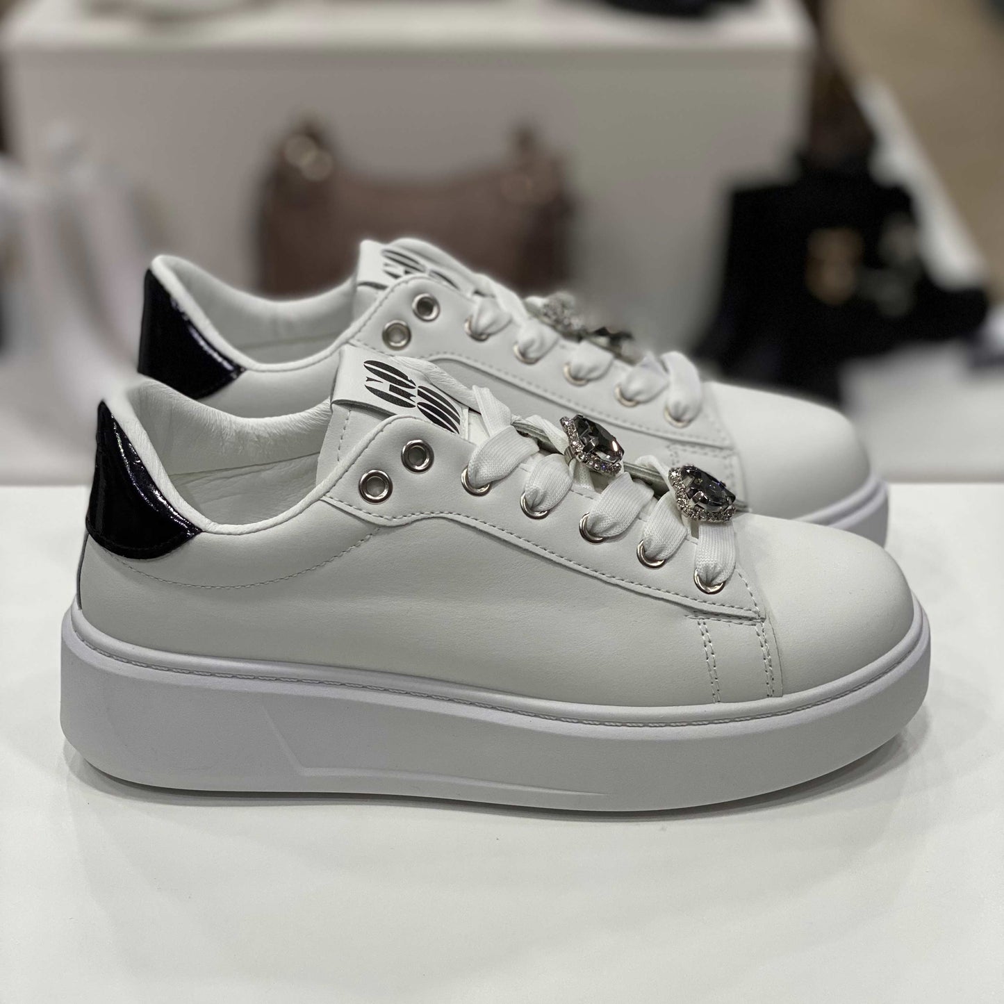 Sneakers bianche con decorazioni gioiello sui lacci e tallone nero lucido, vista laterale su sfondo neutro