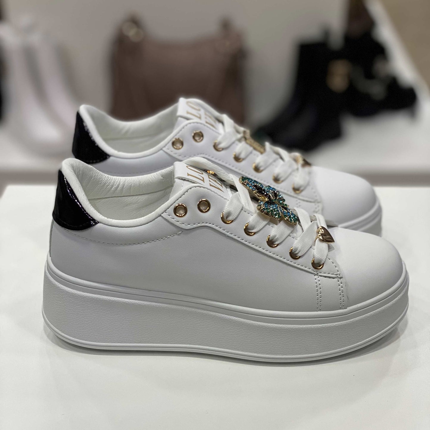 Sneakers bianche con decorazione gioiello a forma di farfalla e dettagli neri lucidi sul tallone, vista laterale per evidenziare il design elegante