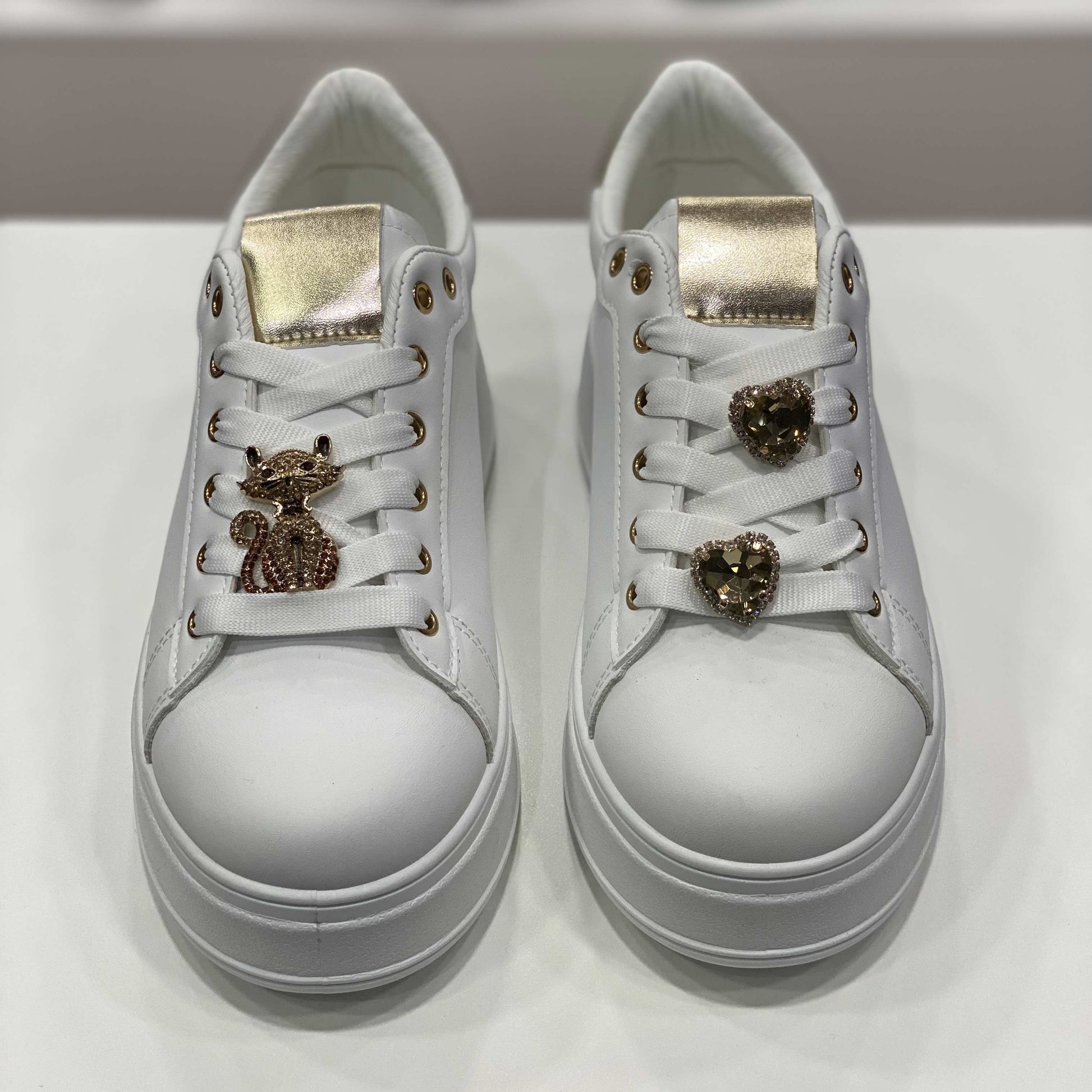 Sneakers bianche con decorazioni gioiello a forma di gatto e cuore, dettagli dorati sulla linguetta e occhielli, vista frontale su sfondo neutro
