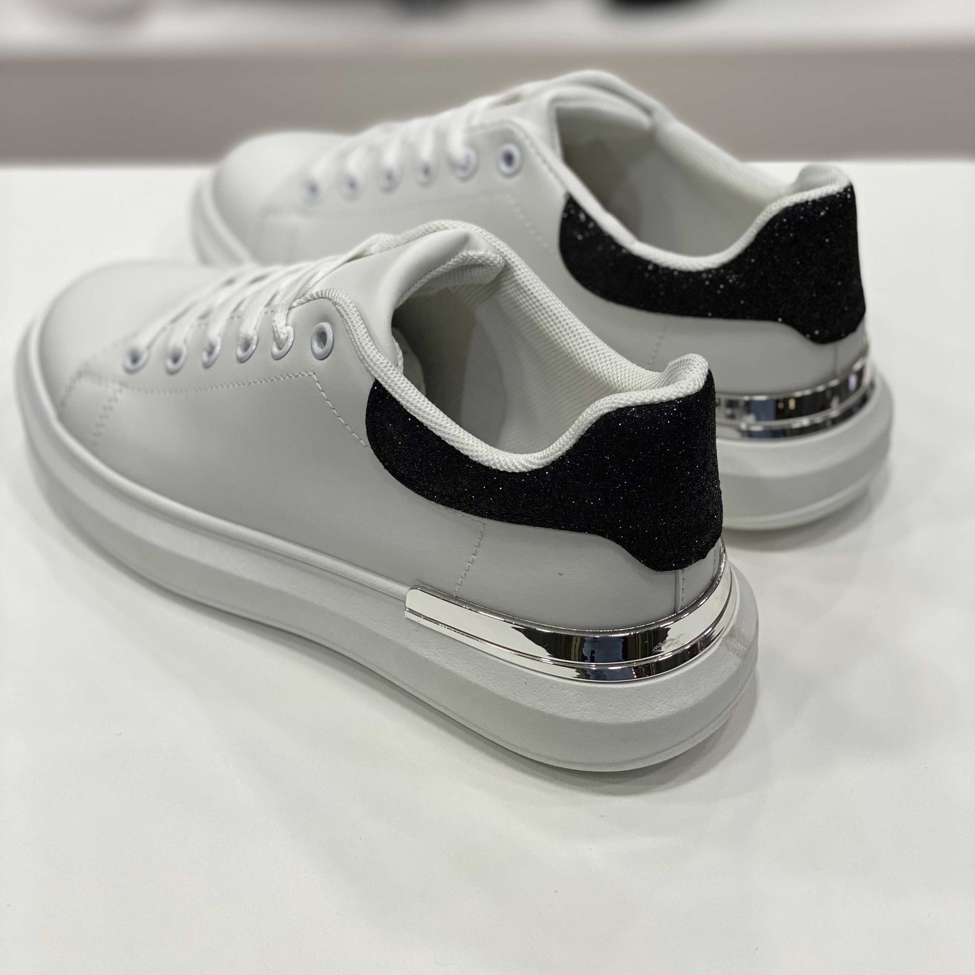 Parte posteriore di sneakers bianche con tallone glitterato nero e rifiniture argentate, vista su sfondo neutro