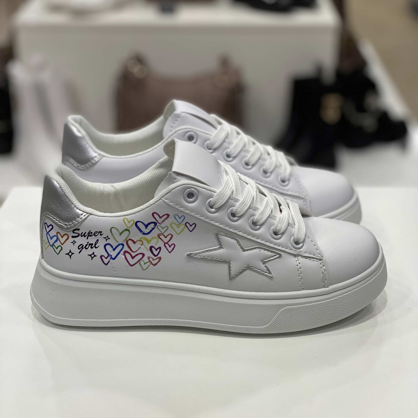 Sneakers bianche con scritta 'Super Girl', cuori colorati e dettaglio a forma di stella e tallone argento, vista laterale su sfondo neutro