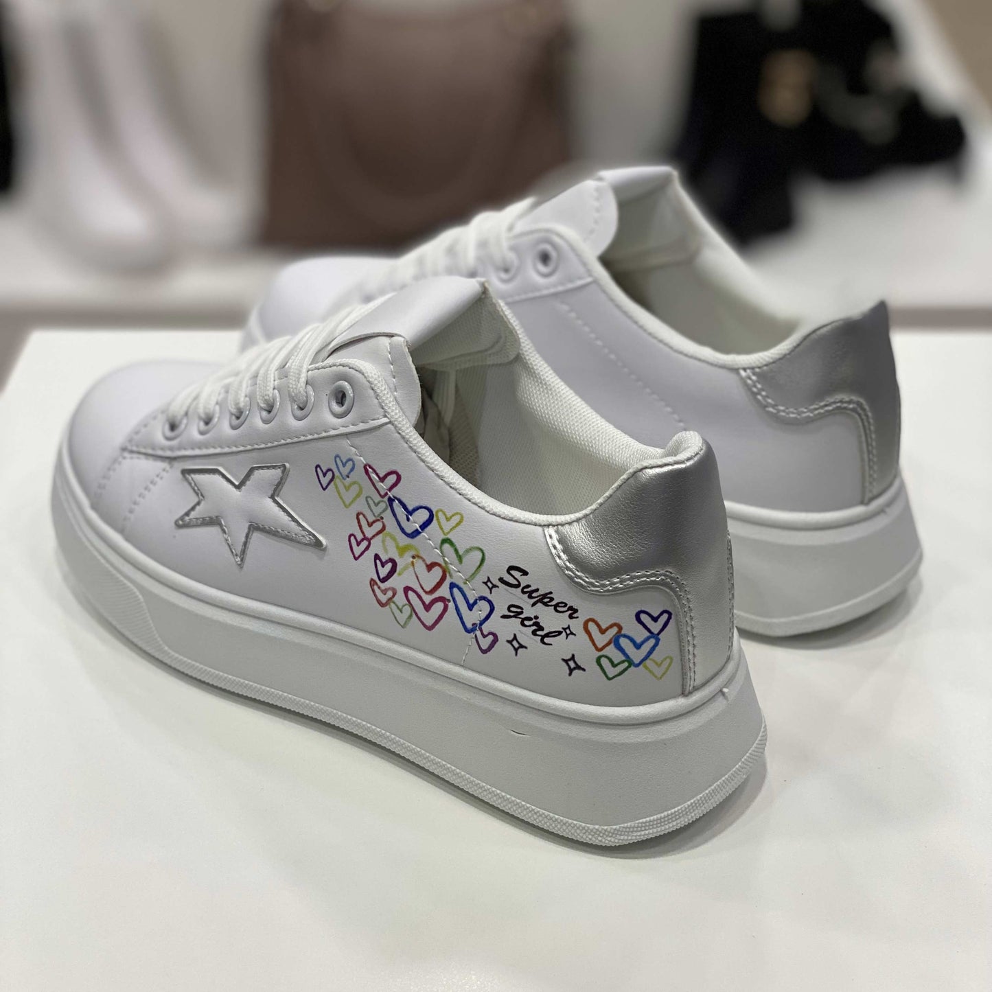 Parte posteriore di sneakers bianche con decorazioni cuori e scritta 'Super Girl', vista del tallone argento su sfondo neutro