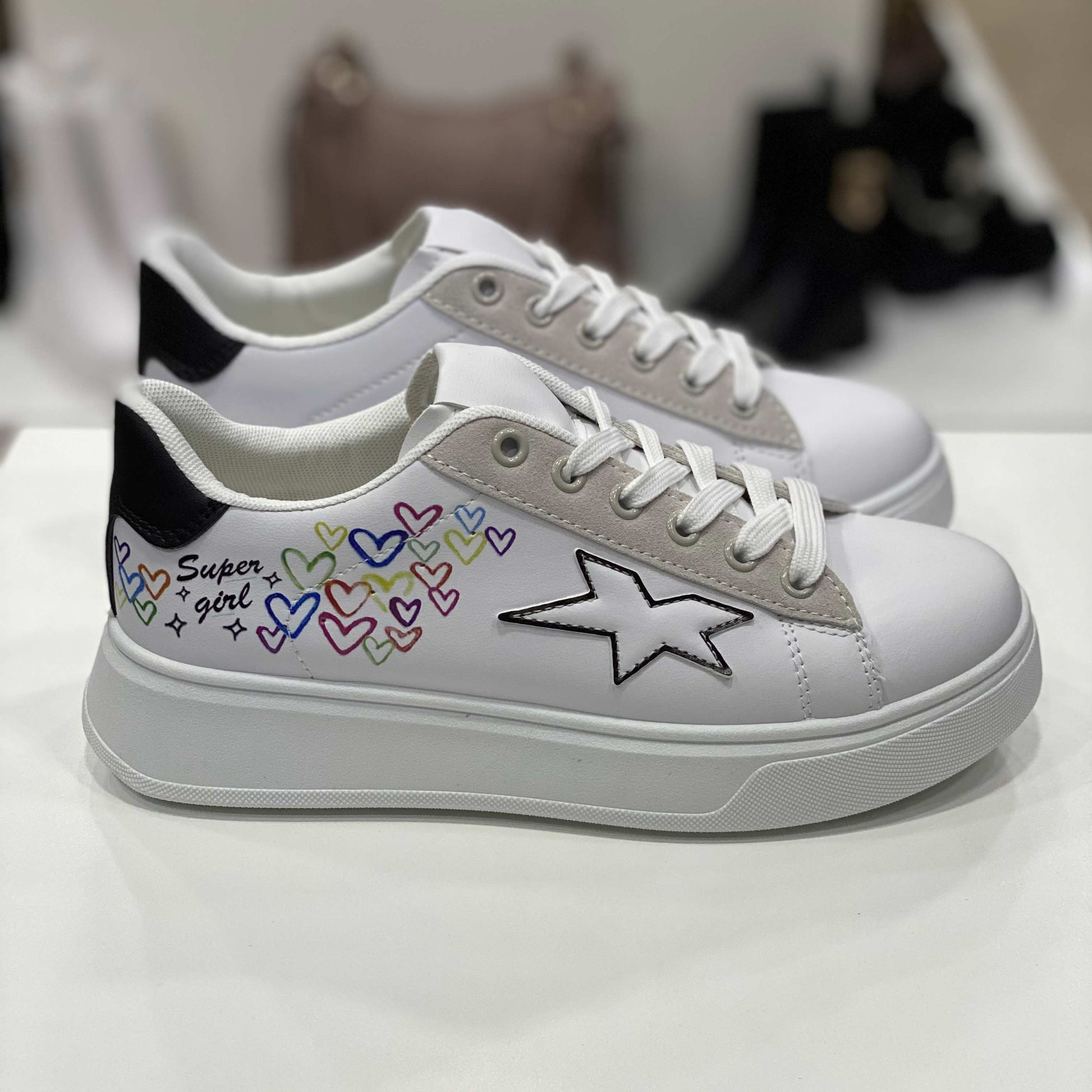 Sneakers bianche con scritta 'Super Girl', cuori colorati e dettaglio a forma di stella e tallone nero, vista laterale su sfondo neutro