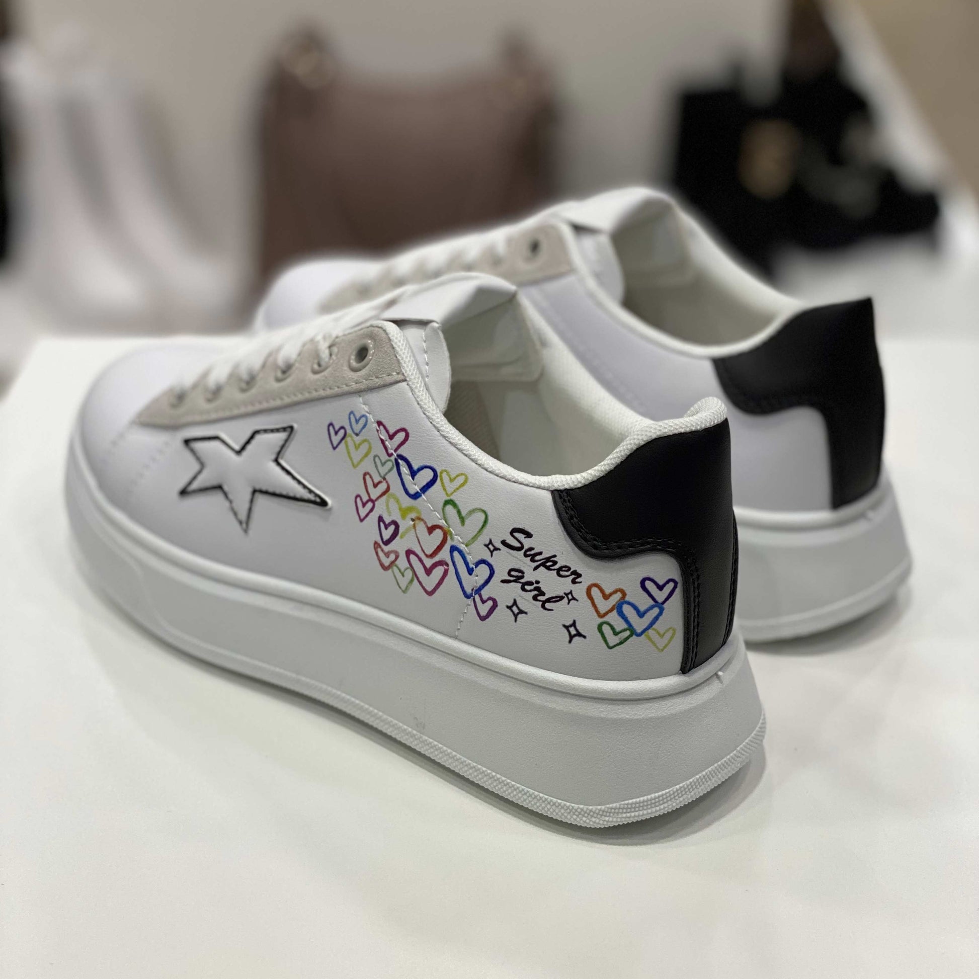 Parte posteriore di sneakers bianche con decorazioni cuori e scritta 'Super Girl', vista del tallone nero su sfondo neutro